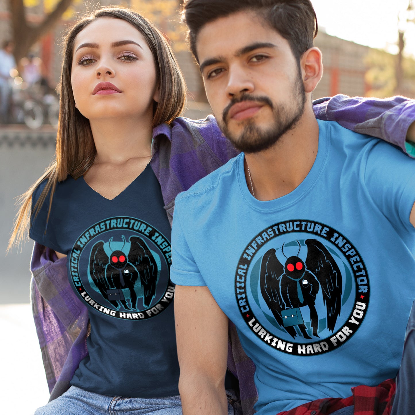 A female model in a navy blue T-shirt and a male model in a light blue T-shirt. Each tee has a black ring/circle printed with the words "Critical infrastructure inspector" in dark blue and "Working hard for you" in white. Within the ring is a black cartoon Mothman with red eyes, a navy tie, and a navy briefcase set against a lighter shadow