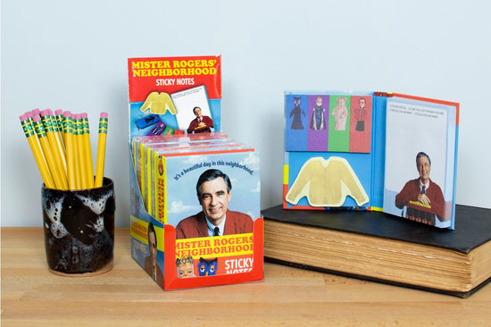 A small book of sticky notes on a wooden desk. The book's cover depicts the Fred Rogers in a red cardigan. Behind him is a blue sky, with "it's a beautiful day in the neighborhood" in blue text. Below him are X the owl and King Friday XIII. Text says "Mister Rogers' Neighborhood sticky notes." Next to the box are sticky notes depicting puppet characters from the series, and a "while you were out" notepad with a picture of Fred Rogers at the bottom. A black jar filled with pencils is next to the notes.