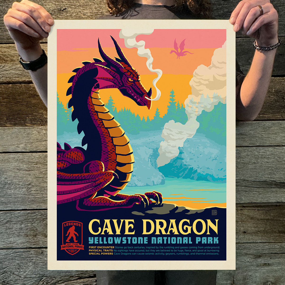 Person holding a poster of a cave dragon in Yellowstone National Park against a wooden background