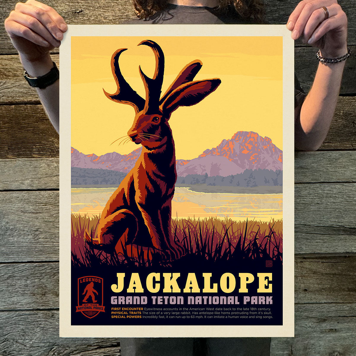 Poster of a jackalope with mountains and water in the background, held by a person on a wooden surface.