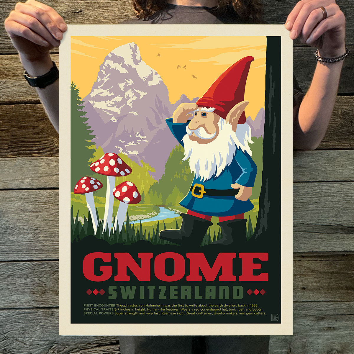 Poster of a gnome in a mountainous landscape held by two hands on a wooden surface.