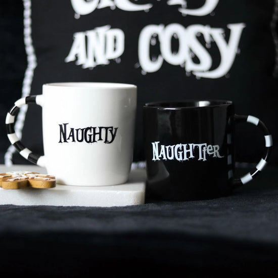 A pair of mugs on a black and white background. The left mug is white with black stripes on the handle. Black text on the mug's center says "naughty." The right mug is black with white stripes on the handle. White text on the mug's center says "naughtier."