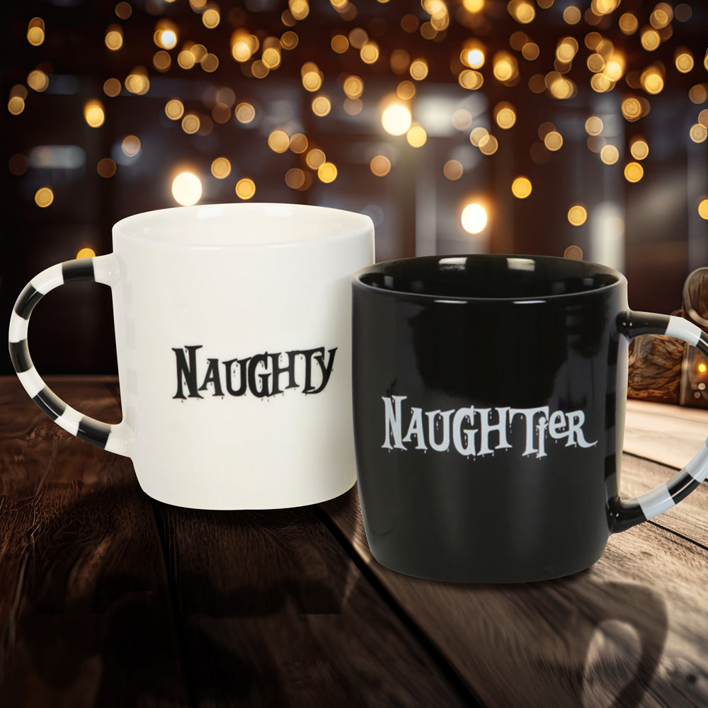 A pair of mugs on a wood table. The left mug is white with black stripes on the handle. Black text on the mug's center says "naughty." The right mug is black with white stripes on the handle. White text on the mug's center says "naughtier." Behind the mugs are a collection of lights hanging from the ceiling of a shadowy room.