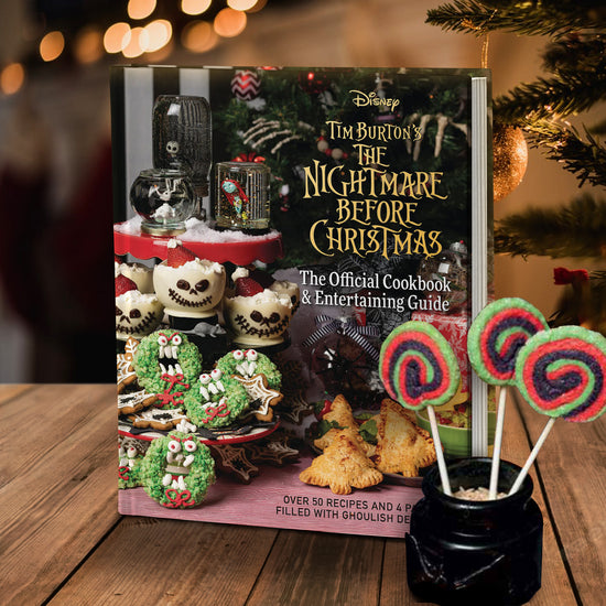 A book on a wooden table, in front of a Christmas tree. On the right side of the cover is yellow text saying "Tim Burton's The Nightmare Before Christmas: The offocial bookbook and entertaining guide." Under that is a tree with halloween-style decorations on the branches. Next to the tree are some of the recipes from the book. A black jar with red, green, and purple swirled cake pops sits beside the book.