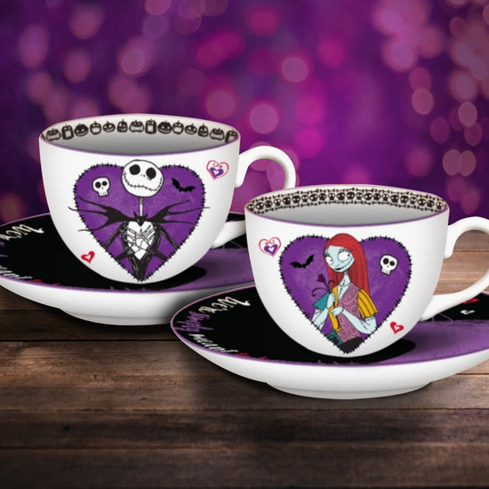 Two bone china teacup and saucer sets on a wood table. Each cup has a purple heart on the front. On one heart is a drawing of Jack Skellington, on the other heart is a drawing of Sally. Each cup comes with a black and purple saucer depicting the two characters standing on the edge of a purple mountain. Behind the cups is a purple background with pink spots.