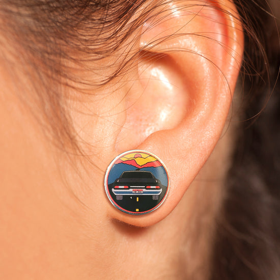 Close up of a model's ear, wearing an earring. The earring depicts a black 1967 Impala driving toward blue mountains, with a red and yellow sky in the distance.