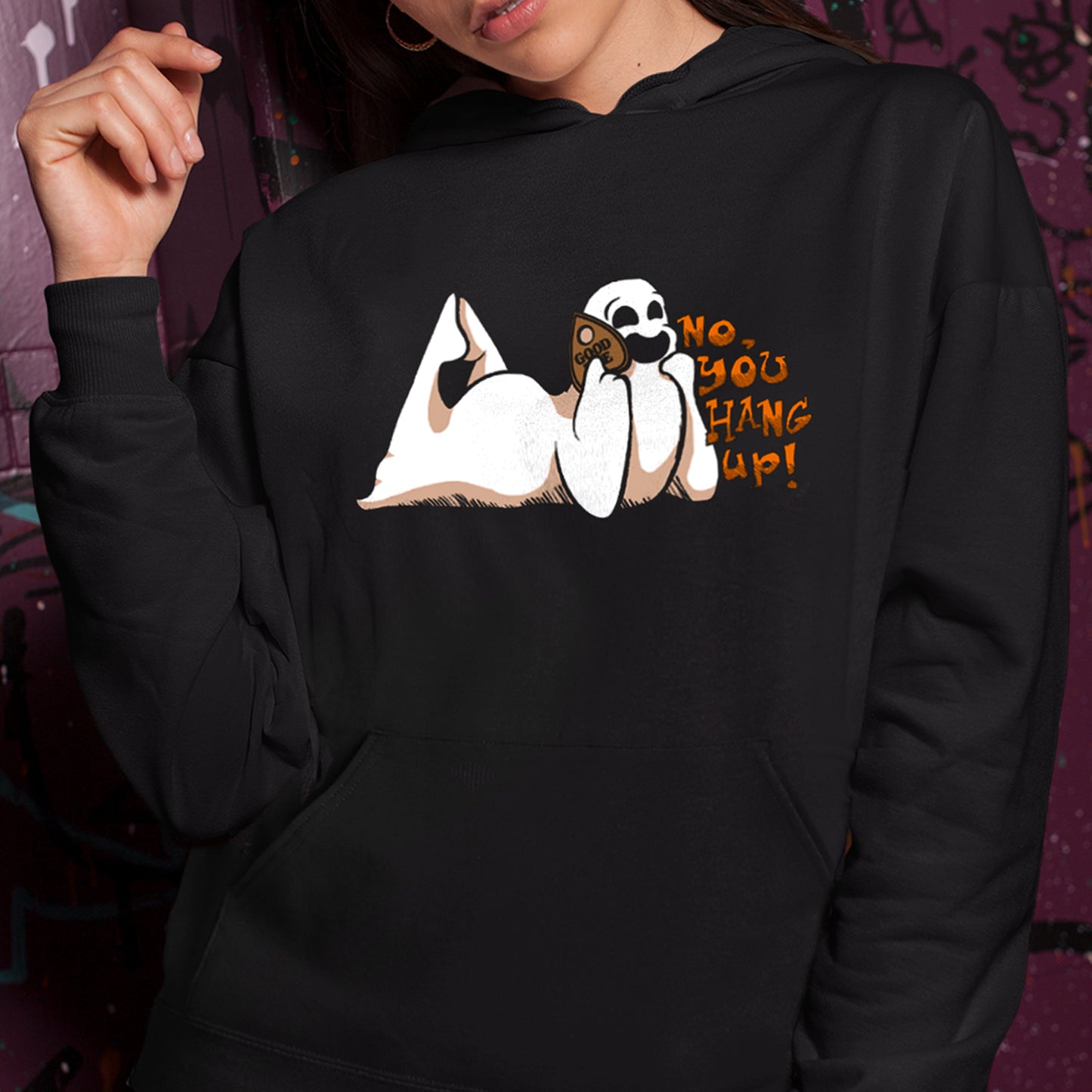 A female model in a black pullover hoodie. The print is a white ghost laying on its stomach with its feet up and a planchette pressed to its ear like a phone. Orange text reads "No, you hang up"