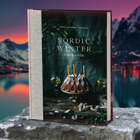 A dark grey book standing on a flat rock. On the cover is a cake sitting on a display plate, with three yellow candles stuck into it. Around the cake are pieces of dark green and grey garland. Behind the cake is a darkly lit forest. Behind the book are dark mountains against a blue, purple, and orange sky, all of which reflect off a lake below them.