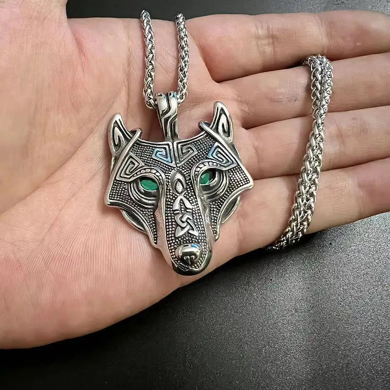 A steel pendant shaped like a wolf's head, laying on a model's open hand. The wolf's eye are made of dark green crystals. The pendant has intricate Nordic style engravings across the surface. At the top is a steel book, attached to a link chain.