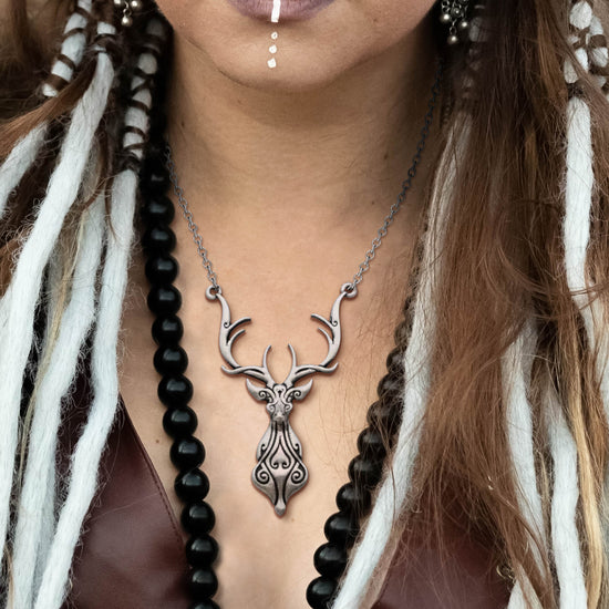 Close up of a silver pendant and chain against a female model's neckline. The pendant is shaped like the head, chest, and antlers of a deer, with black swirls drawn on the deer's curves. The model has long white tassles woven into her brown hair, and is wearing a necklace of black beads.