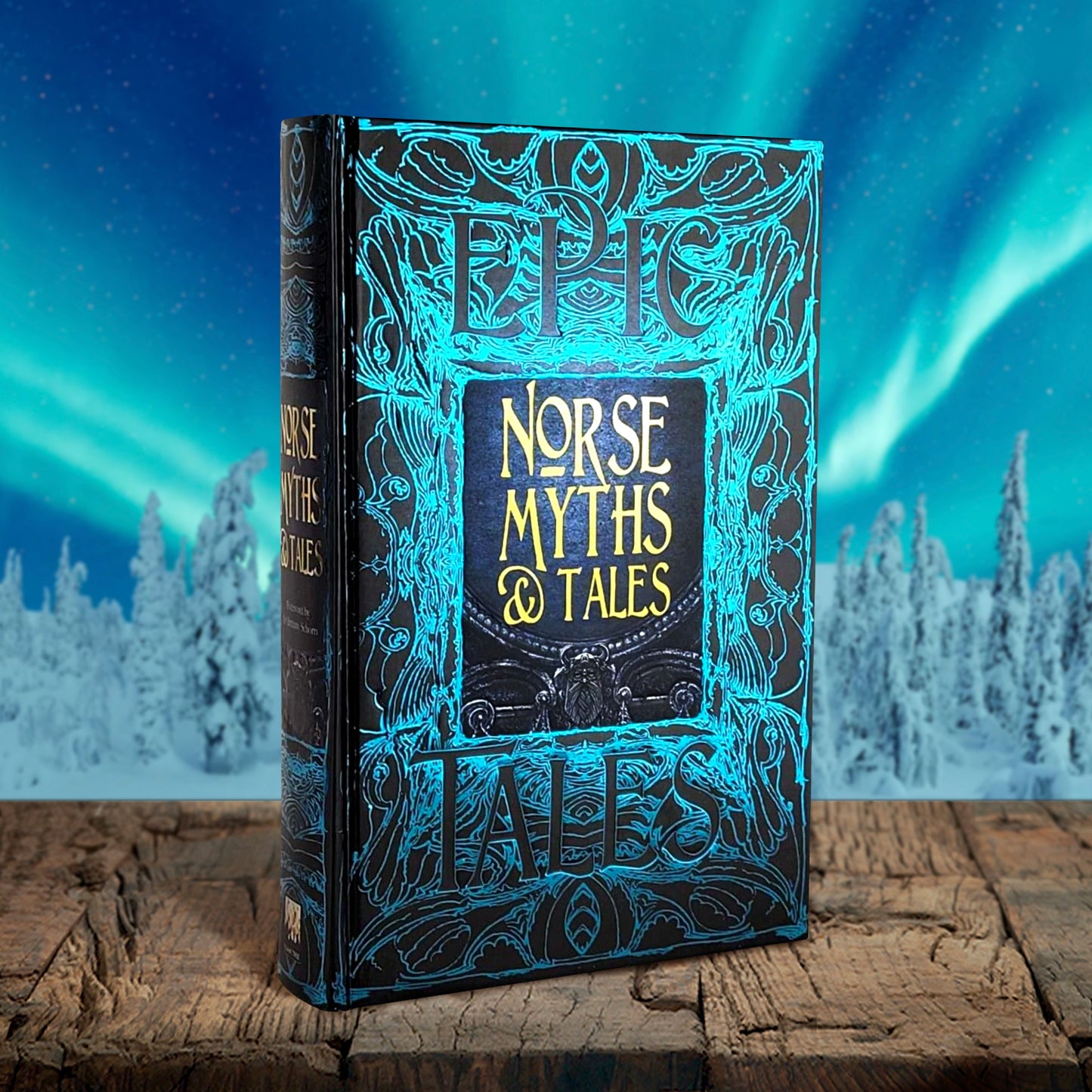A blue book on a wood table. The background of the cover depicts blue and black patterns, similar to tree bark. At the center is a black square with yellow text saying "norse myths & tales." At the top and bottom is black text saying "epic tales." Behind the book is a winter forest with the Northern Lights visible in the sky.