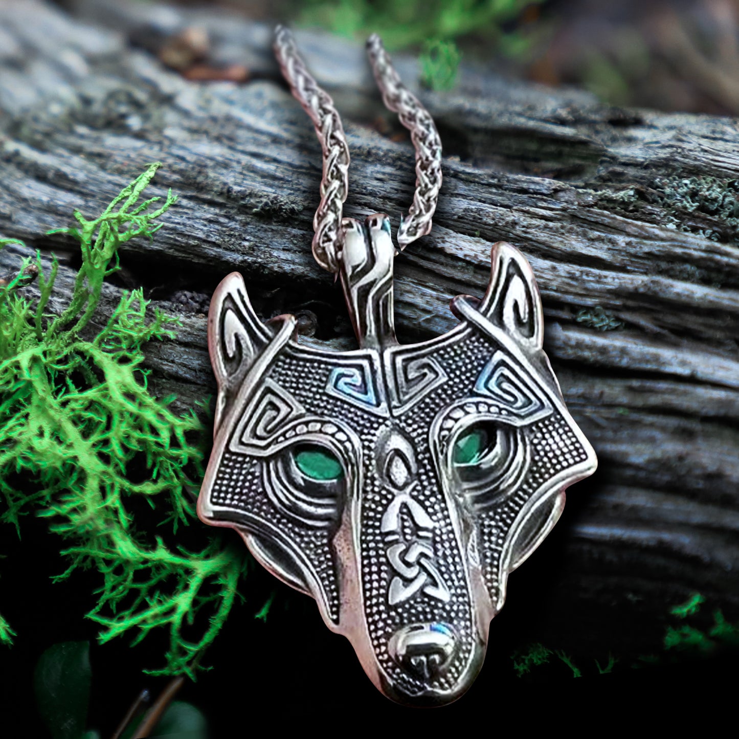 A steel pendant shaped like a wolf's head, laying on a dark grey tree limb. The wolf's eye are made of dark green crystals. The pendant has intricate Nordic style engravings across the surface. At the top is a steel book, attached to a link chain. Next to the pendant is a sprig of green moss.