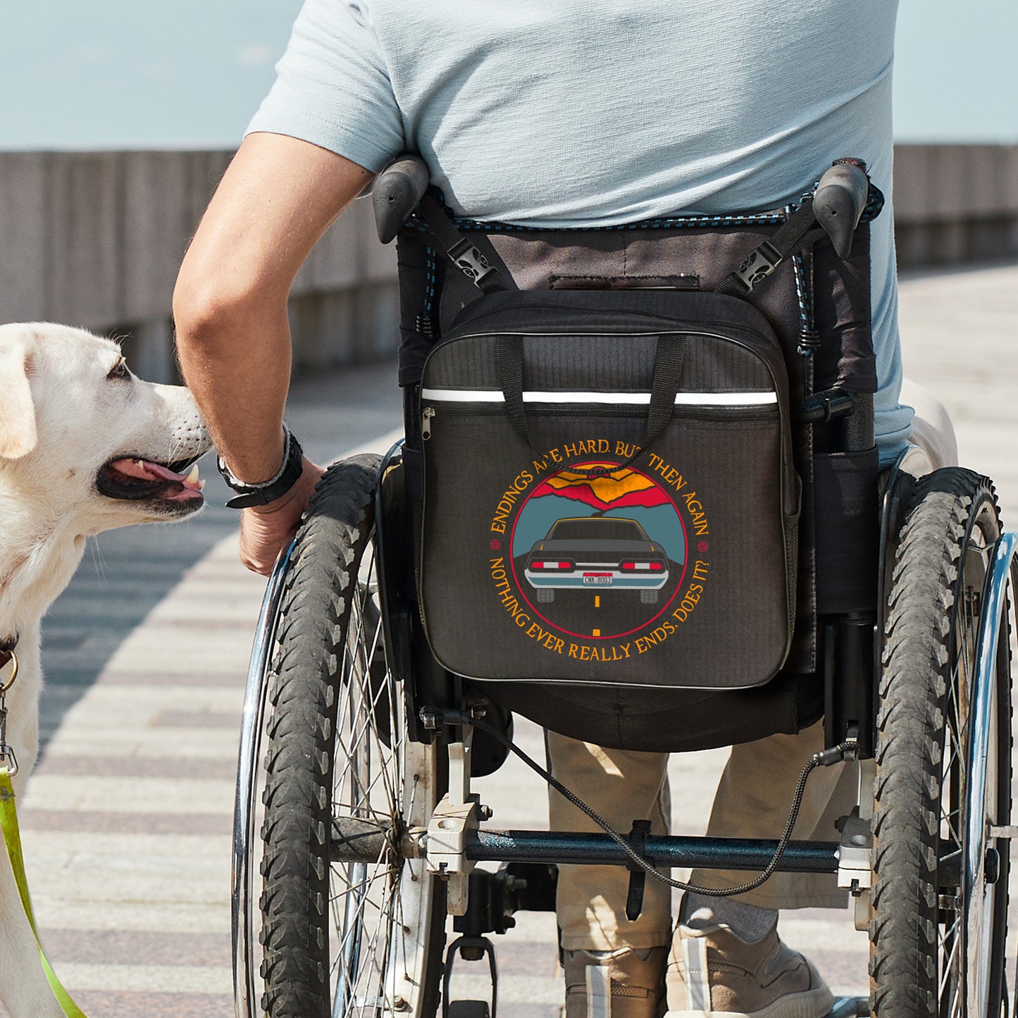 An image of a model in a wheelchair. On the back of the wheelchair is a black messenger-style bag. The bag depicts the tail end of a black 1967 Chevy Impala driving on a black road toward a sunset. Around the edge is yellow text saying “Endings are hard, but then again nothing ever really ends, does it?" A blonde dog sits next to the model.