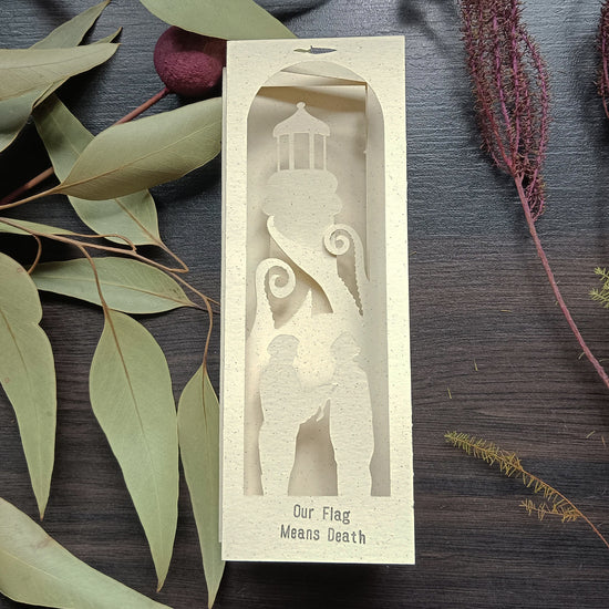 A white paper bookmark against a dark wood background, with leaves and sprigs around it. The bookmark depicts a lighthouse, and silhouette cut-outs of characters from the series "Our flag means death." In front of the lighthouse are kraken tentacles. Silver text at the bottom of the bookmark says "our flag means death."