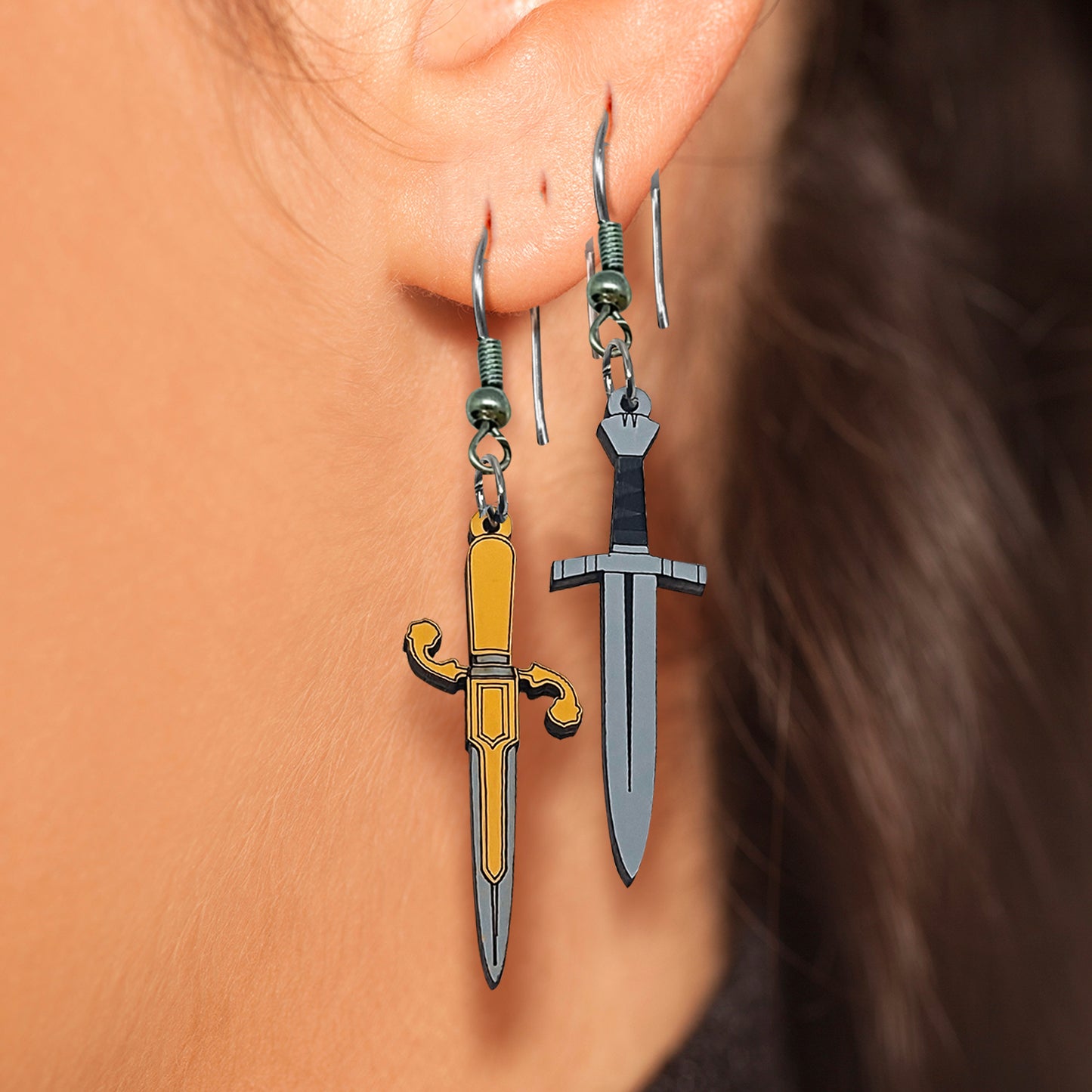 Close up view of a model's ear. Two dagger-shaped earrings hang from her earlobe. One dagger is gold with an asymmetrical hilt. The other is black and grey with a straight hilt.