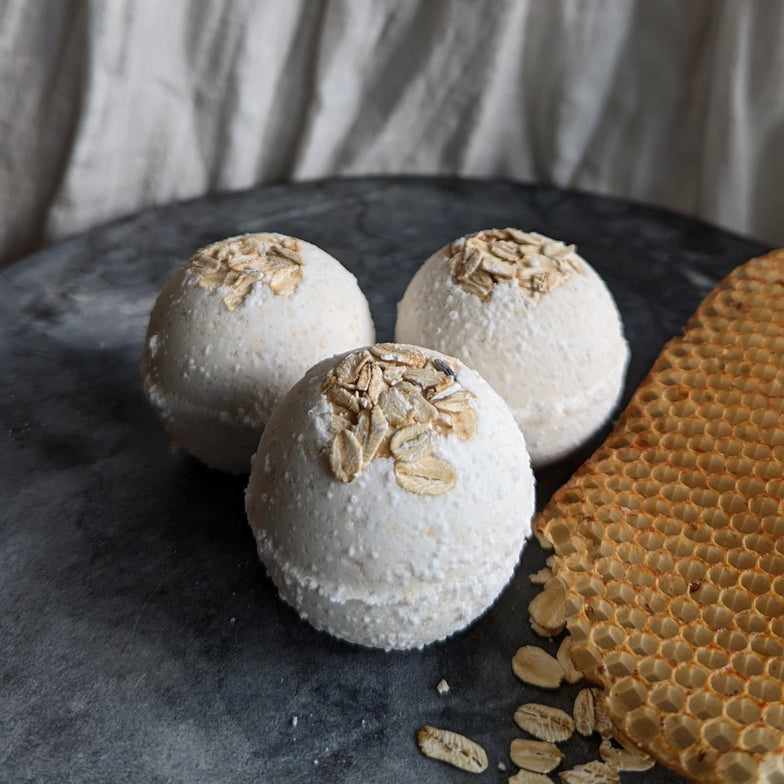 Close up of three white bath bombs on a dark stone table. The top of each bath bomb is covered in chopped oats. Next to the bath bombs is a piece of natural honeycomb. Behind the table is a white curtain.