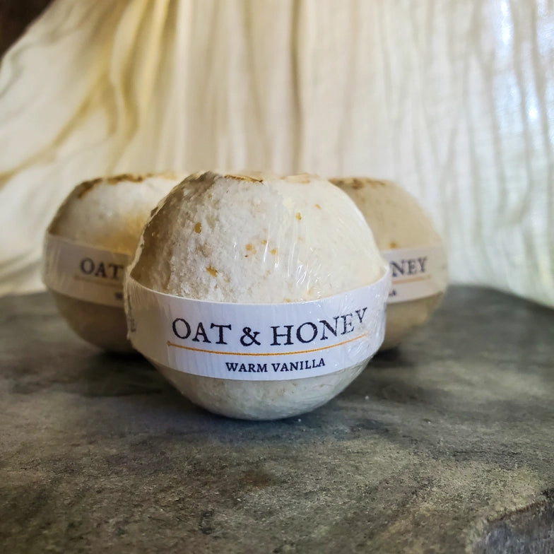 Close up of three white bath bombs on a dark stone table. Each bath bomb is wrapped in plastic, with a label across the front. On the label is dark blue text that says "Oat and honey, warm vanilla." Behind the bath bombs is a white curtain.