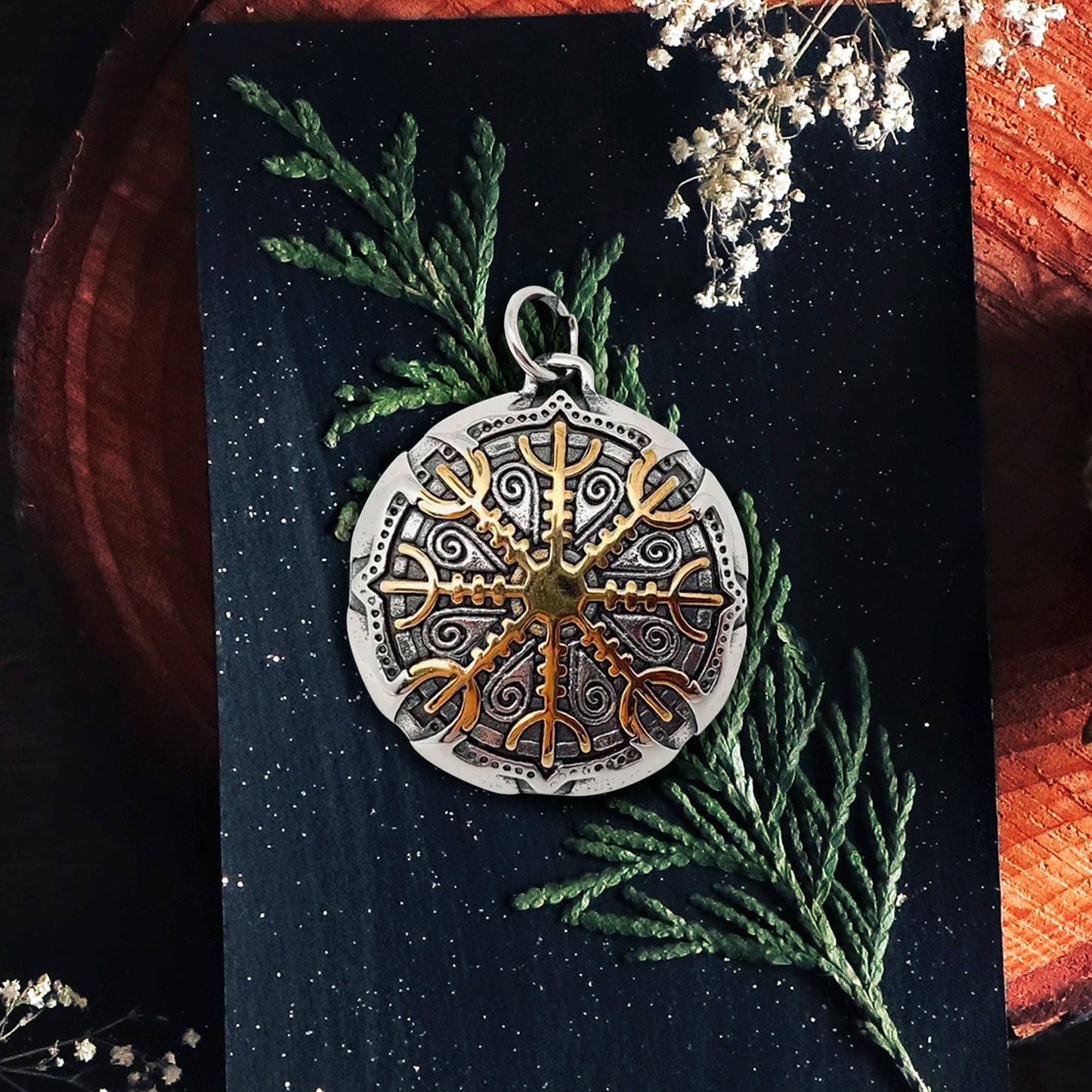 A stainless steel pendant decorated with Nordic-style runes, against a blue cloth speckled with white dots. The front has eight trident-shaped sections, plated in gold. At the top of the pendant is a metal loop, to hand on a chain. Behind the pendant is a sprig of green herbs. In the background is the flat circle of a cut tree trunk.