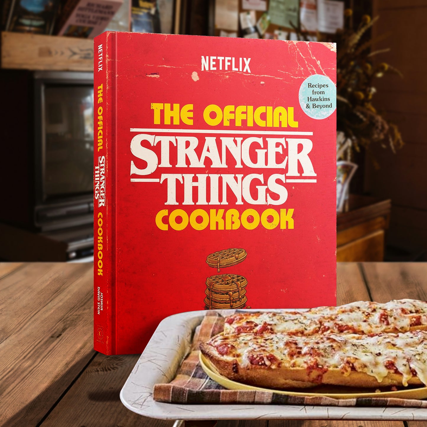 Red 'The Official Stranger Things Cookbook' on a table with pizza slices.