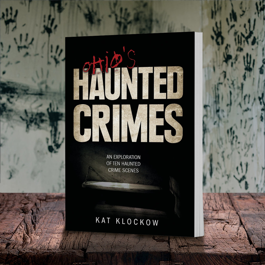 An image of a black book on a wooden table, against a wall covered in handprints. At the center of the cover in white and red text is "Ohio's Haunted Crimes: an exploration of ten haunted crime scenes." Under the text is a dark and grainy image of a prison cell cot.