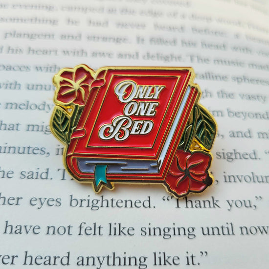 A red enamel pin on a book page. The pin is shaped like a book, with roses on each side, White text on the cover says "only one bed."