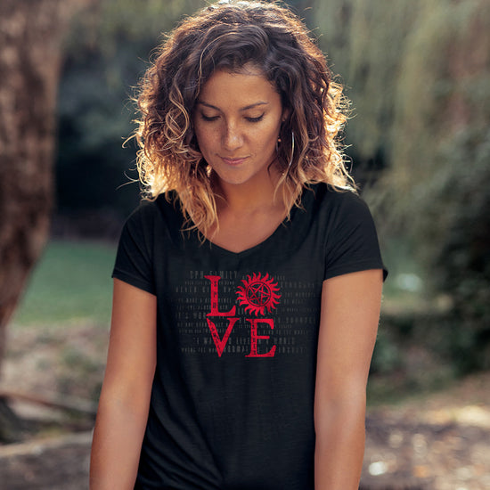 A female model wearing a black v-neck t-shirt with short sleeves and red lettering that stacks LOVE - the "o" is the anti-possession symbol. Behind the model is a forest.