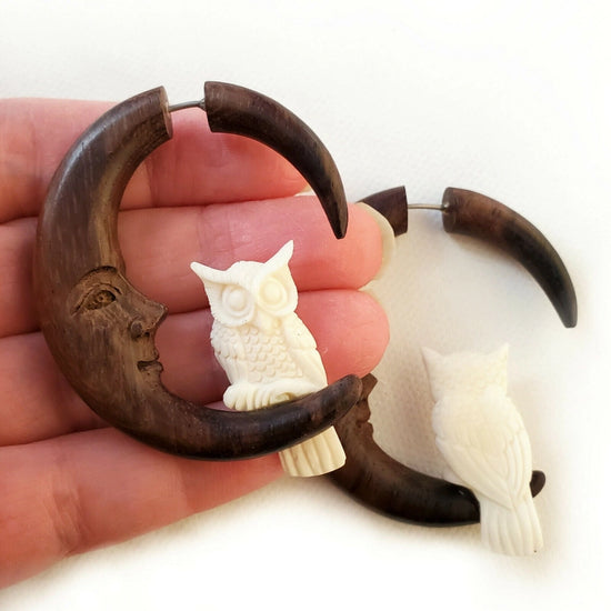 Front and back image of a pair of wooden earrings shaped like a crescent moon. At the bottom of each is a small white owl. One earring sits on a model's hand, the other is against a white background. The inside of the crescent moon shows a smiling face