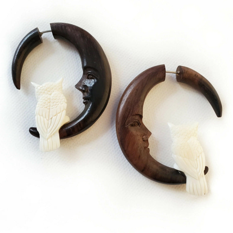 Two wooden earrings side by side on a white background. Each earring is shaped like a crescent moon, with a small white owl at the bottom. The inside of the crescent moon shows a smiling face.