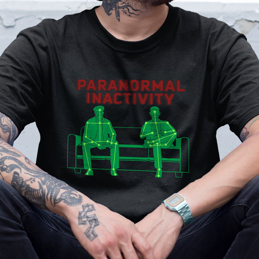 A model in a black tee shirt. On the front is a green drawing of two men on a couch, with lines and dot representing their shapes. Red text says paranormal inactivity"