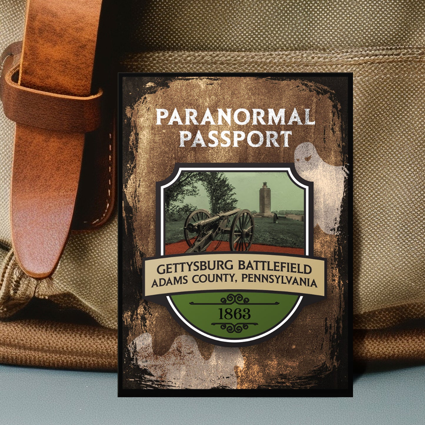 Close up on an enamel pin. The pin has a drawing of Civil War battlefield in the center, surrounded by a stone-colored background. Two white ghost drawings are in the background. At the top is white text saying "Paranormal passport." Under the battlefield drawing is text saying "Gettysburg Battlefield. Adams County, Pennsylvania 1863."