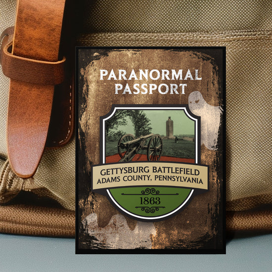 Close up on an enamel pin. The pin has a drawing of Civil War battlefield in the center, surrounded by a stone-colored background. Two white ghost drawings are in the background. At the top is white text saying "Paranormal passport." Under the battlefield drawing is text saying "Gettysburg Battlefield. Adams County, Pennsylvania 1863."