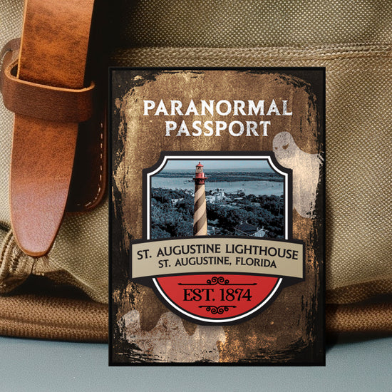 Close up on an enamel pin. The pin has a drawing of striped lighthouse on the shore in the center, surrounded by a stone-colored background. Two white ghost drawings are in the background. At the top is white text saying "Paranormal passport." Under the battlefield drawing is text saying "St. Augustine Lighthouse, St. Augustine Florida. 1874."