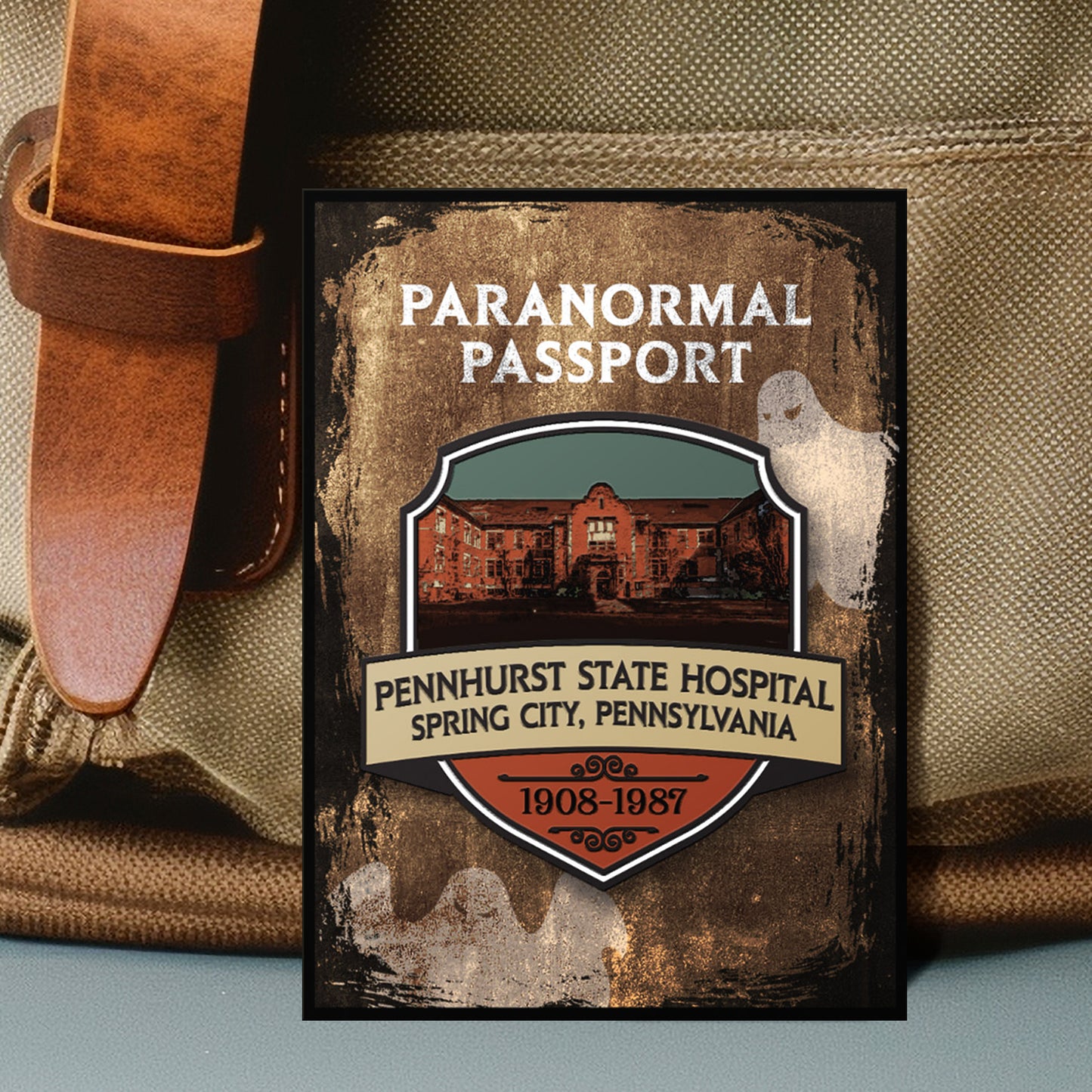 Close up on an enamel pin. The pin has a drawing of an old hospital in the center, surrounded by a stone-colored background. Two white ghost drawings are in the background. At the top is white text saying "Paranormal passport." Under the hospital drawing is text saying "pennhurst state hospital, spring city, pennsylvania. 1908-1987."