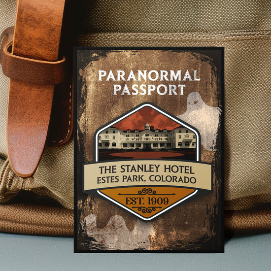 Close up on an enamel pin. The pin has a drawing of an old hotel in the center, surrounded by a stone-colored background. Two white ghost drawings are in the background. At the top is white text saying "Paranormal passport." Under the hotel drawing is text saying "The Stanley Hotel. Estes Park, Colorado Est 1909."