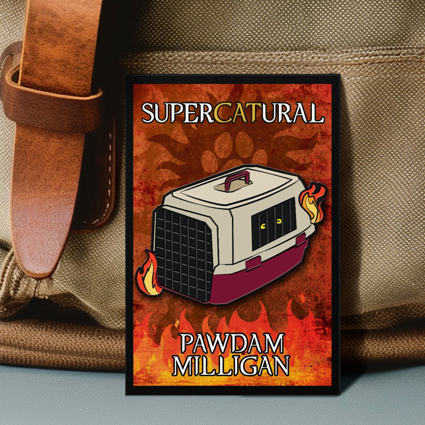 A red and orange enamel pin against a canvas backpack. The pin depicts a cartoon drawing of a cat carrier surrounded by fire. Visible in one of the carrier's caged openings are two yellow cat eyes. White and yellow text says "supercatural: pawdam milligan." The background of the pin is a reddish orange with a black paw print, reminiscent of the anti-possession symbol.