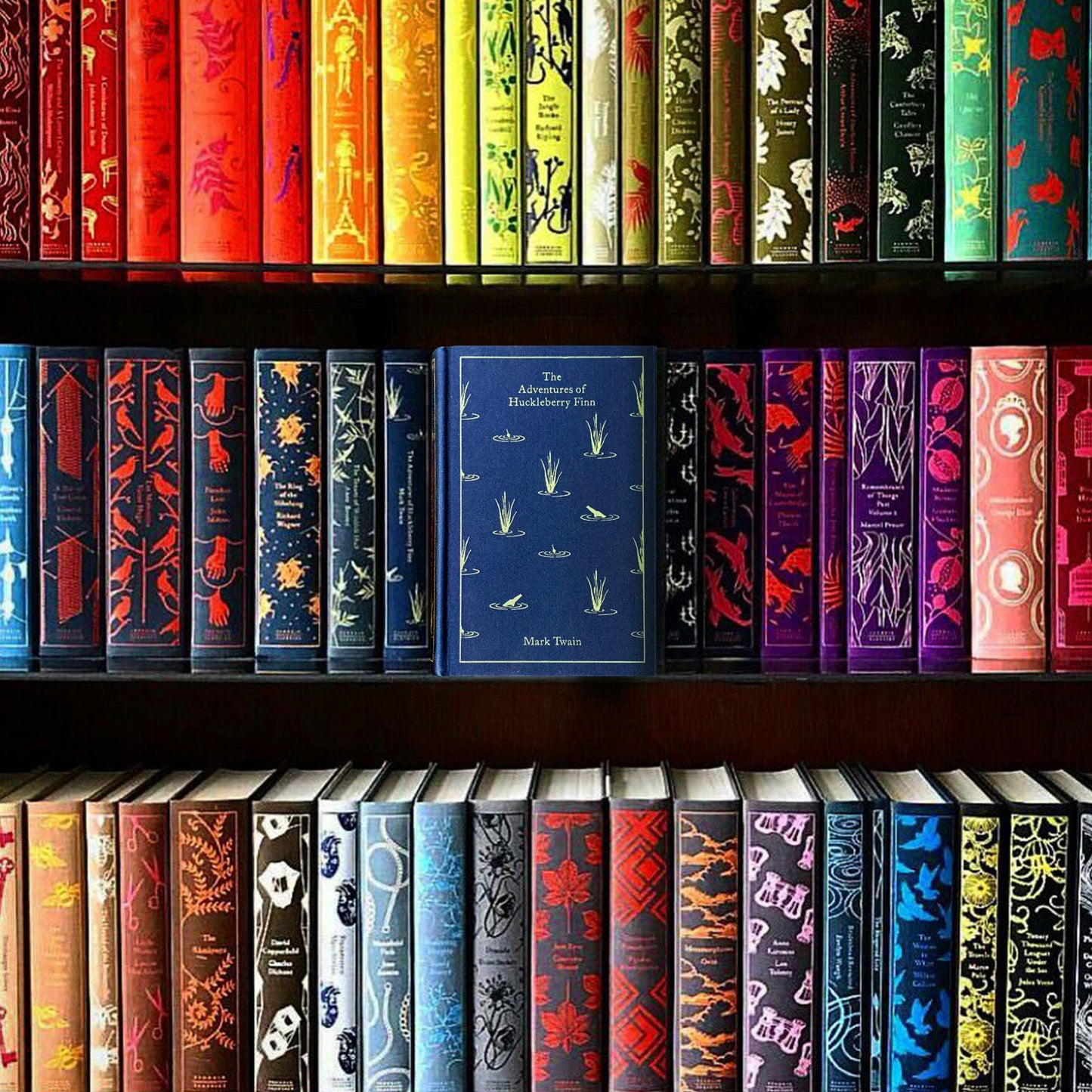 A collection of colorful books lined up on three bookshelves. In the center is a blue book with yellow text saying "The adventures of huckleberry finn, Mark Twain."