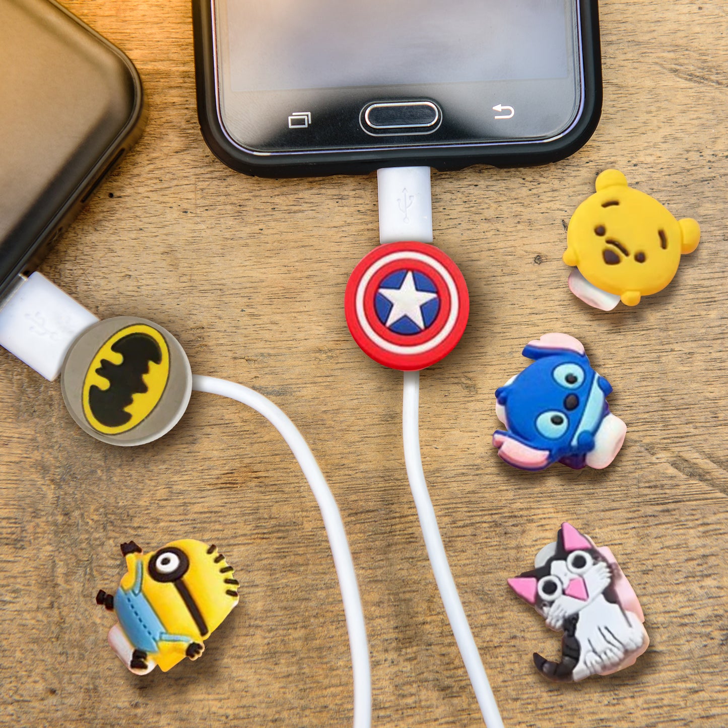 Close up view of two cell phones connected to charger cables, on a wooden background. Attached to the charger cables are plastic grippers. One depicts Captain America's shield, the other depicts Batman's symbol. Next to the phones are four more grippers in varying designs: Pooh bear, a Minion, Stitch, and a black and white cat.