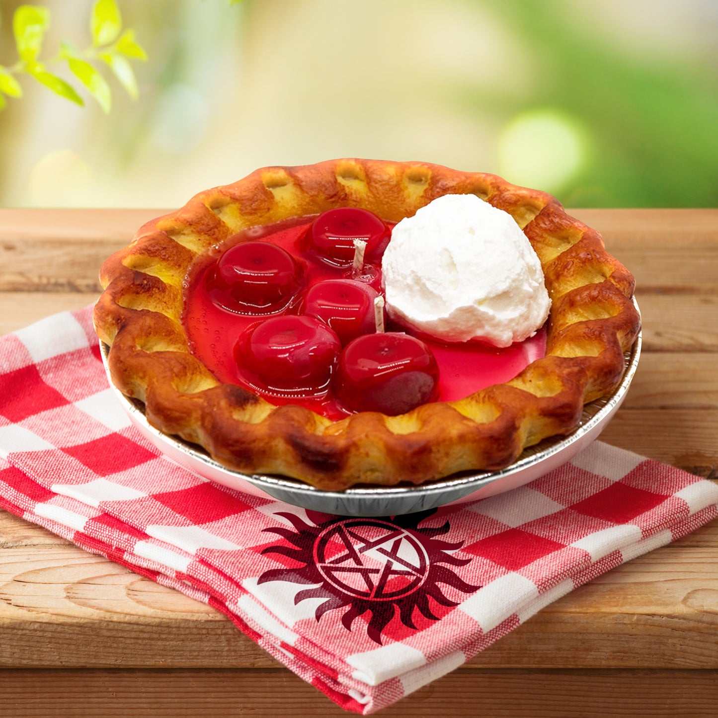 Close up view of a candle shaped like a cherry pie with whipped cream, sitting on a wooden table. Under the candle is a red and white checkered napkin with the anti-possession symbol on one corner.