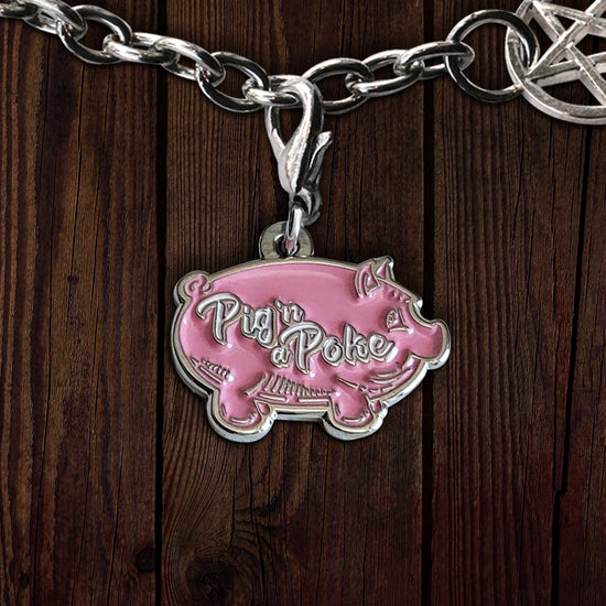 A pink pig-shaped charm on a silver chain, against a dark wooden background. Across the middle of the pig is white scripted text that says "pig n a poke.