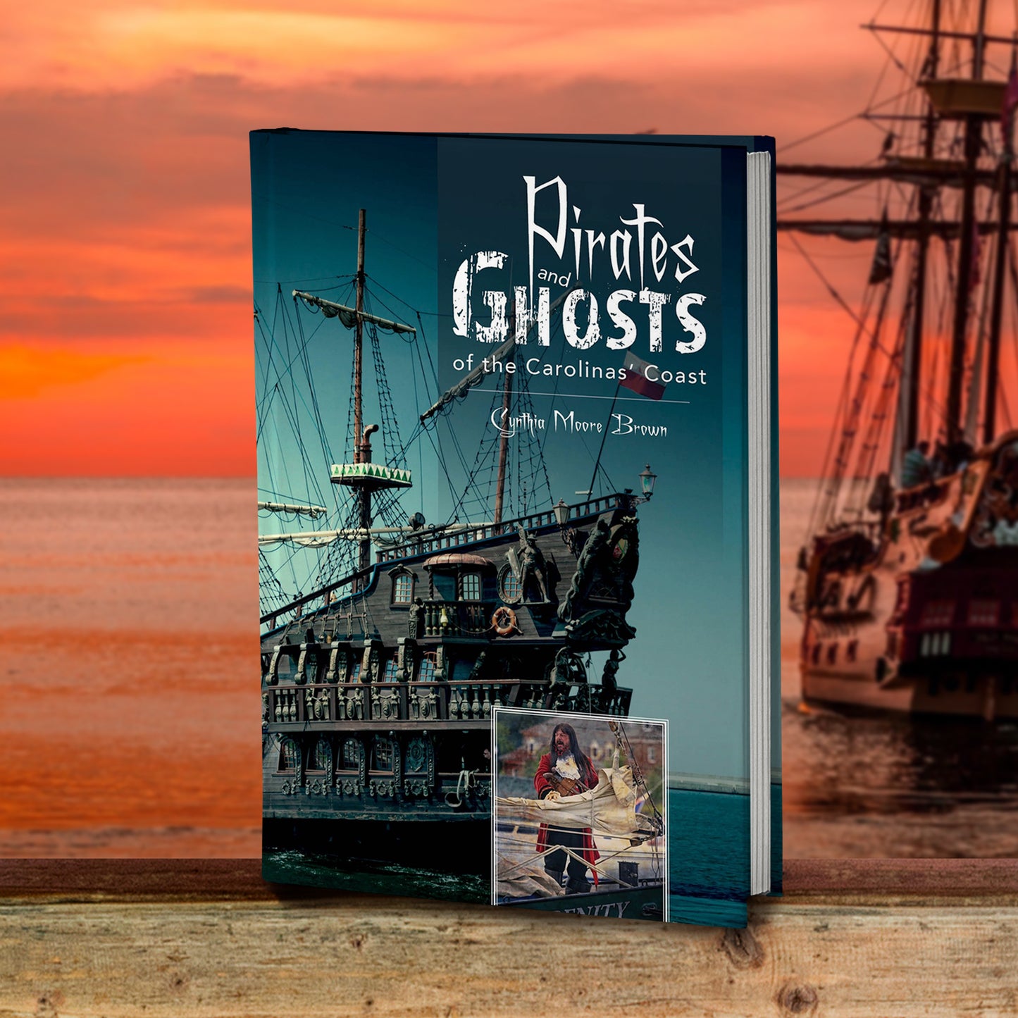An image of a book standing on a beach, with a sunset sky above it. On the right is a wooden ship. On the cover is old pirate ship on the sea. At the top of the cover in white text is "Pirates and ghosts of the carolina's coast." At the bottom of the cover is an inset image of a man dressed in pirate clothes, standing in front of a folded sail.