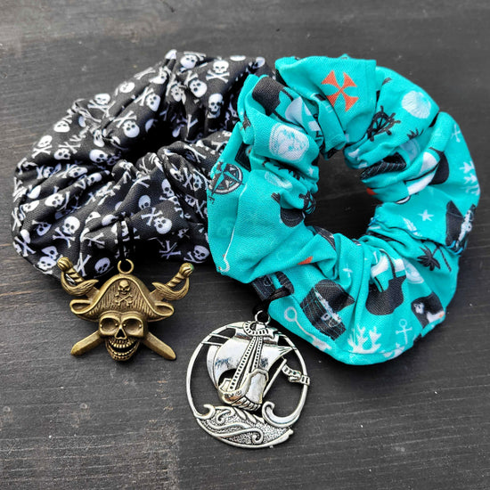 A pair of scrunchies on a black table. One scrunchie is black with a white skull and crossbones pattern. Attached to the scrunchie is a brass charm, shaped like a skull and crossbones with a pirate hat. The second scrunchie is light blue, with drawings of pirate ships and other pirate-themed items. A silver charm is attached to the bottom of the scrunchie, depicting a pirate ship on the sea.