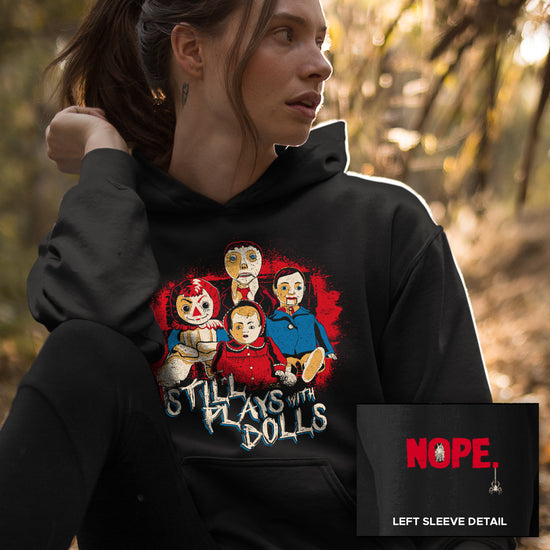 A female model wearing a black hoodie against a white background. On the shirt is a collection of creepy dolls, surrounded by red spatters. Under the dolls is white text that says "still plays with dolls." At the bottom corner is a close up of the sleeve, with red text saying "NOPE" and a small spider hanging from the last letter.