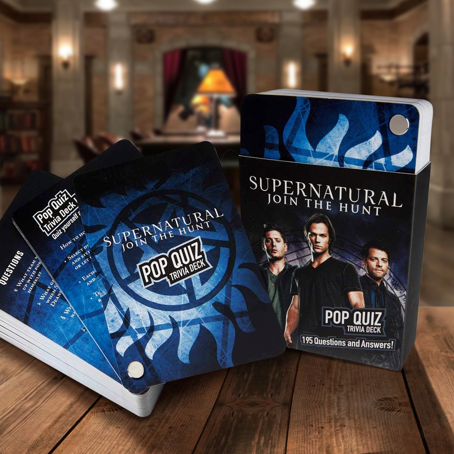 A deck of blue and black trivia cards. Around the deck is a black banner with images of Sam and Dean Winchester and Castiel on the bottom. At the top is white text saying "Supernatural: join the hunt." At the bottom is white text saying Pop quick trivia deck, 195 questions and answers! The deck is sitting on a wooden table in front of the Men of Letters Bunker set from Supernatural.