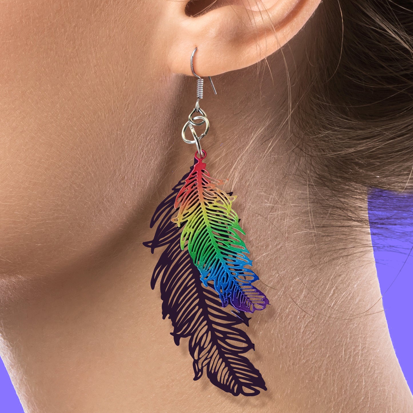 Close up of a model wearing a dangle earring. The earring is shaped like two feathers of different sizes. The smaller feather is rainbow colored, the larger feather is a dark purple.