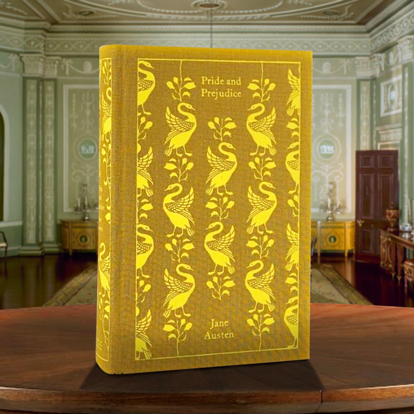 A yellow and gold book on a wooden table. The cover of the books depicts swans and lilies. Gold text at the top and bottom says "Pride and prejudice, Jane Austen." Behind the book is a sitting room in a mansion from the early 1800s.