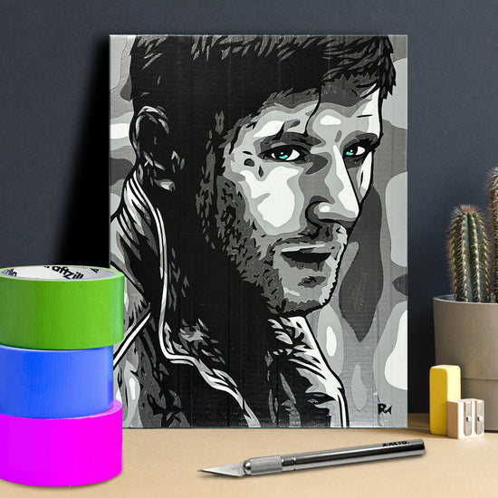 A black and white poster against a dark gray wall. The poster depicts Dean Winchester, created through shades of duct tape. Next to the poster are rolls of colored duct tape and cutting tools.