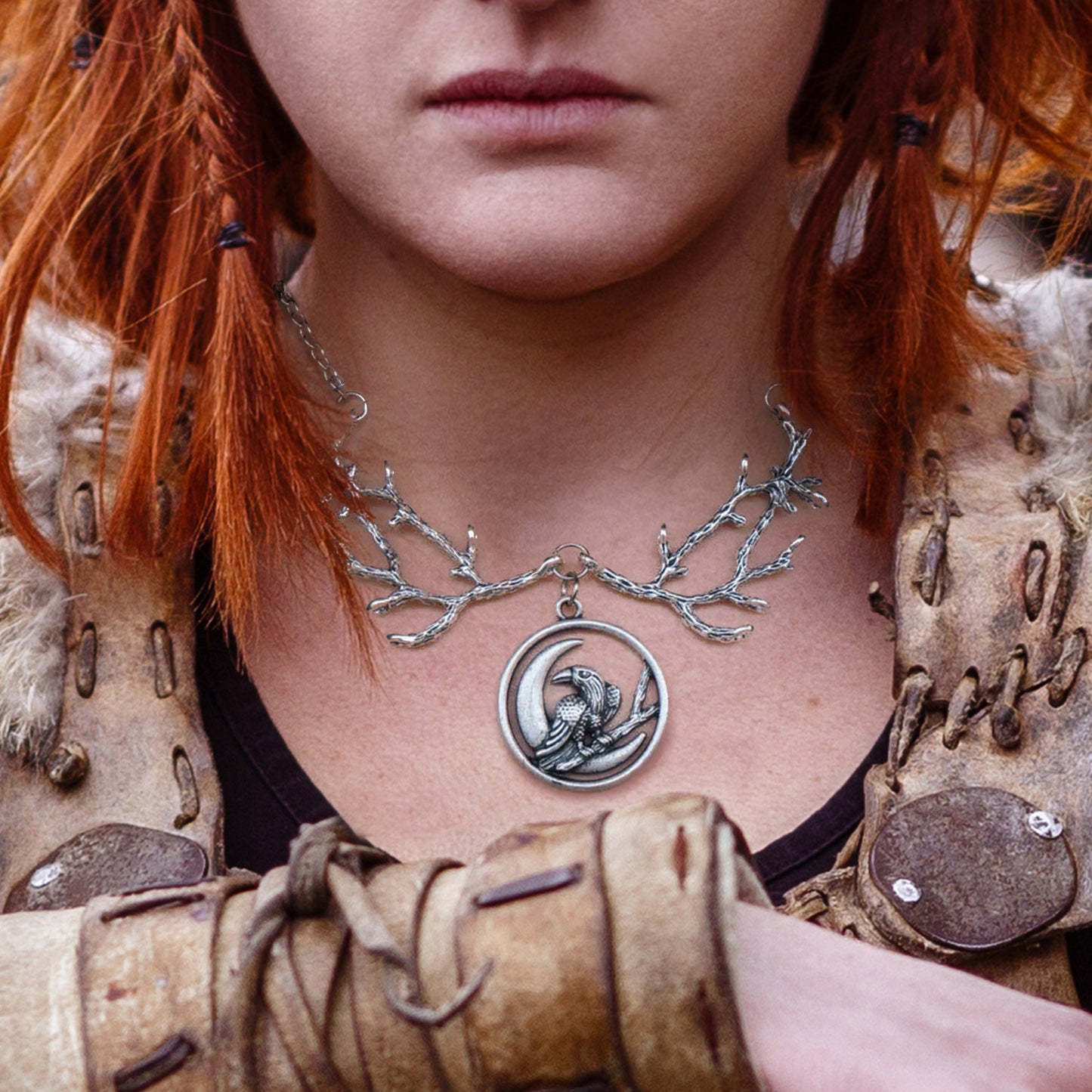 Close up of a red-haired female model wearing a silver necklace. The necklace has an upper section in the shape of tree branches. A round pendant hangs from the bottom of the branches. Inside the pendant is a crow perched on a twig, in front of a crescent moon. The model is dressed in Viking-style clothing.
