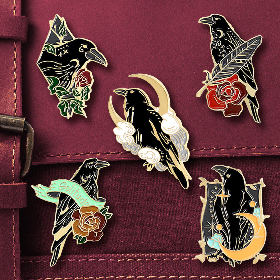 Five enamel pins attached to a maroon messenger bag. Each pin depicts a black raven.