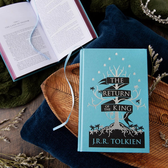 Two blue hardcover books on a wooden tray. The front cover of one book depicts a minimalist drawing of a white tree on a black ground. White text says "The Return of the King." The other book is open to the start of a chapter.