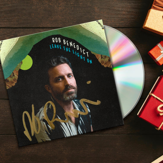 A picture of a CD case featuring a photo of Rob Benedict and the title "Leave the Lights On". The case is signed by Rob in gold Sharpie and a CD is poking out from the sleeve. The CD is sitting on a wooden table and there are several small, red and gold gifts sitting on the table next to it.